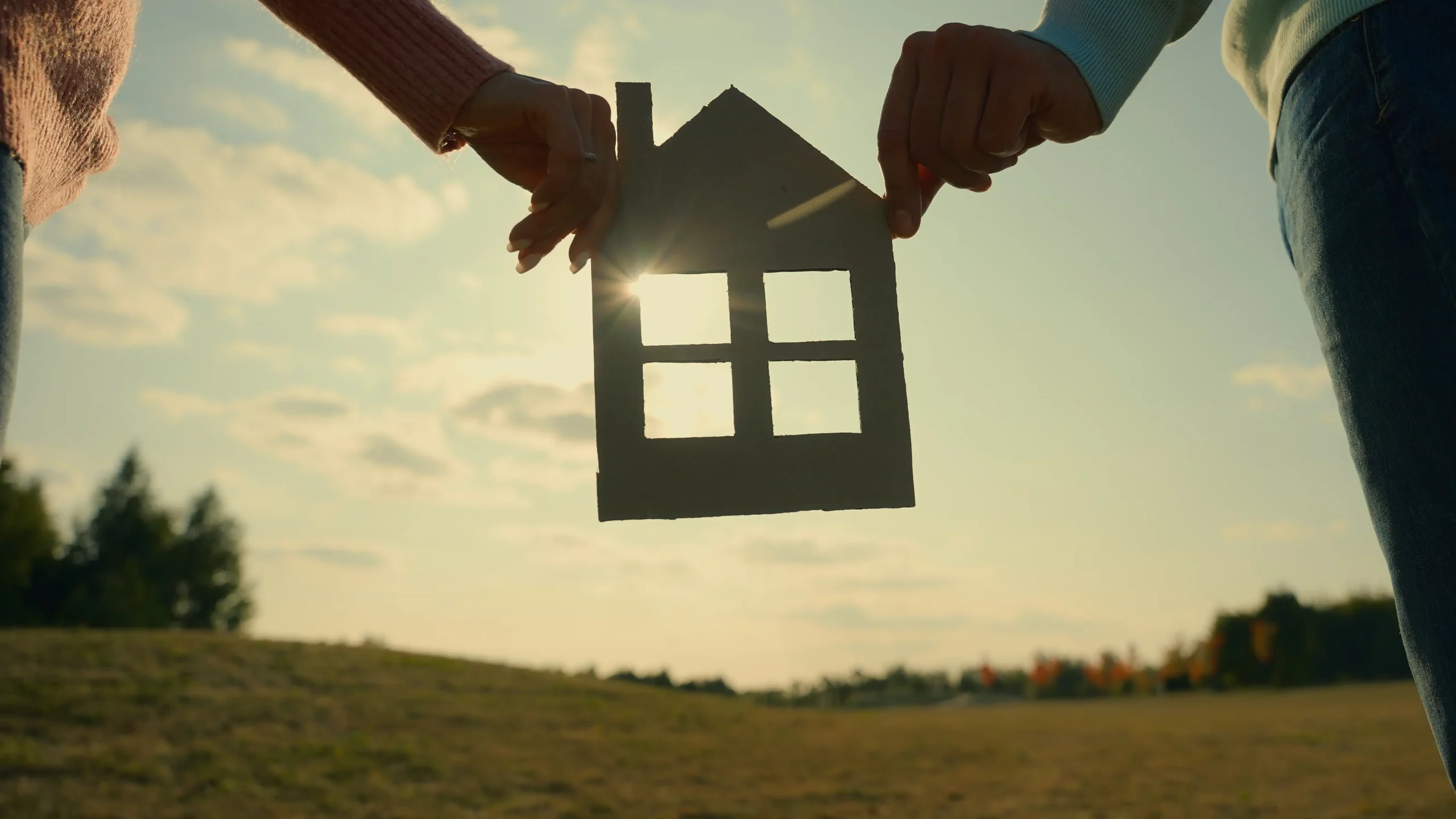 Zwei Hände halten ein Modellhaus vor eine Wiese im Sonnenuntergang
